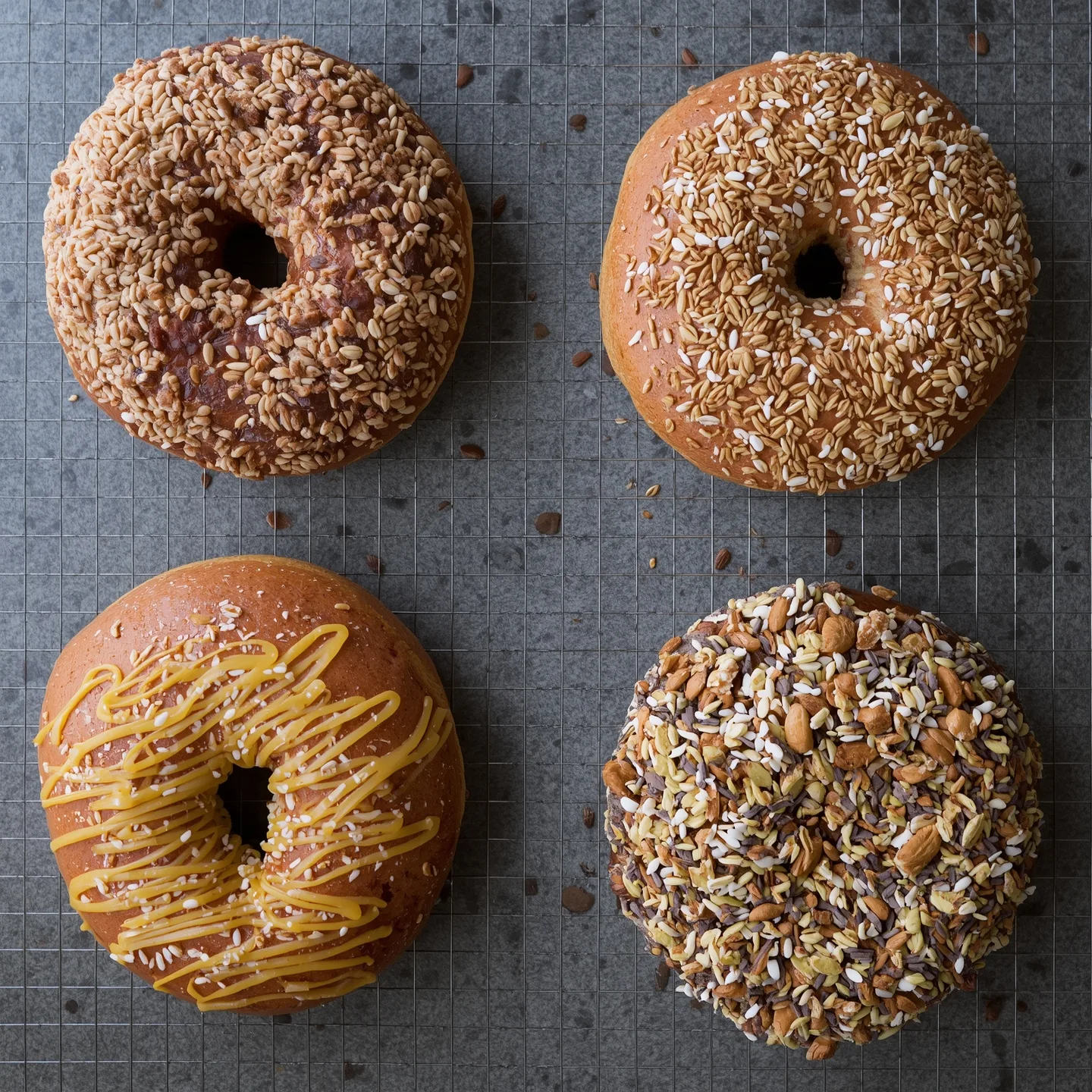 Four freshly baked bagels, each with a different topping including sunflower seeds, a sweet drizzle, and a mix of seeds and almonds, arranged on a cooling rack.