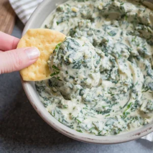 A hand dipping a cracker into a creamy bowl of spinach dip with a thick, rich texture.