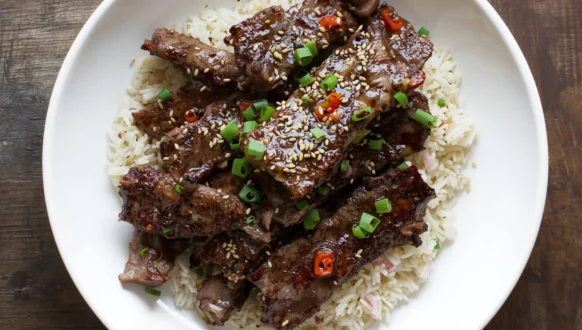 Plate of glossy Korean beef recipe over white rice, sprinkled with sesame seeds, chopped green onions, and small red chili pieces.