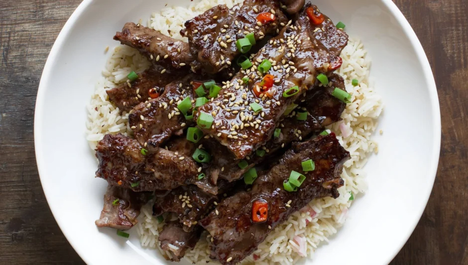 Plate of glossy Korean beef recipe over white rice, sprinkled with sesame seeds, chopped green onions, and small red chili pieces.